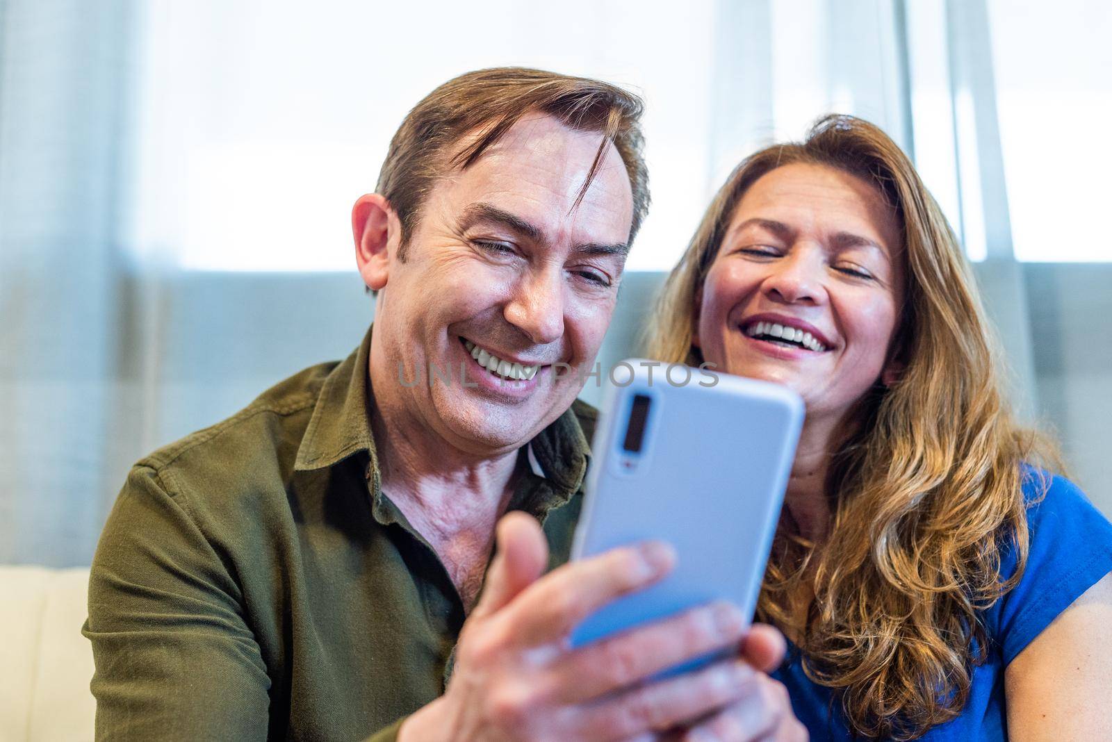 Smiling mature couple at home watching something funny on mobile. by ivanmoreno