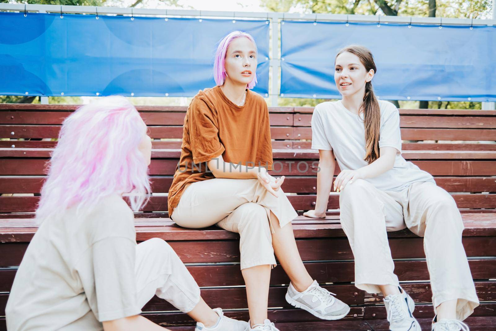 Modern teenage girls with colorful dyed hair sitting on bench in park. Women chatting, gossiping and laughing. Friendship concept. by kristina_kokhanova