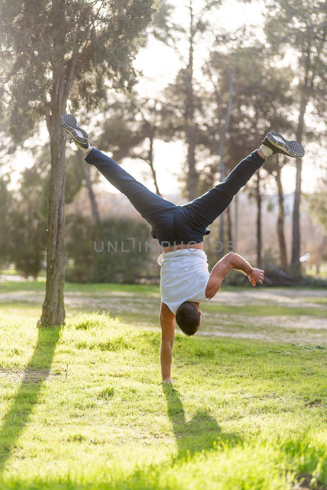 Gymnastic man handstand on one hand doing acrobatic posture. Athletic man wearing white tank top doing handstand in park.