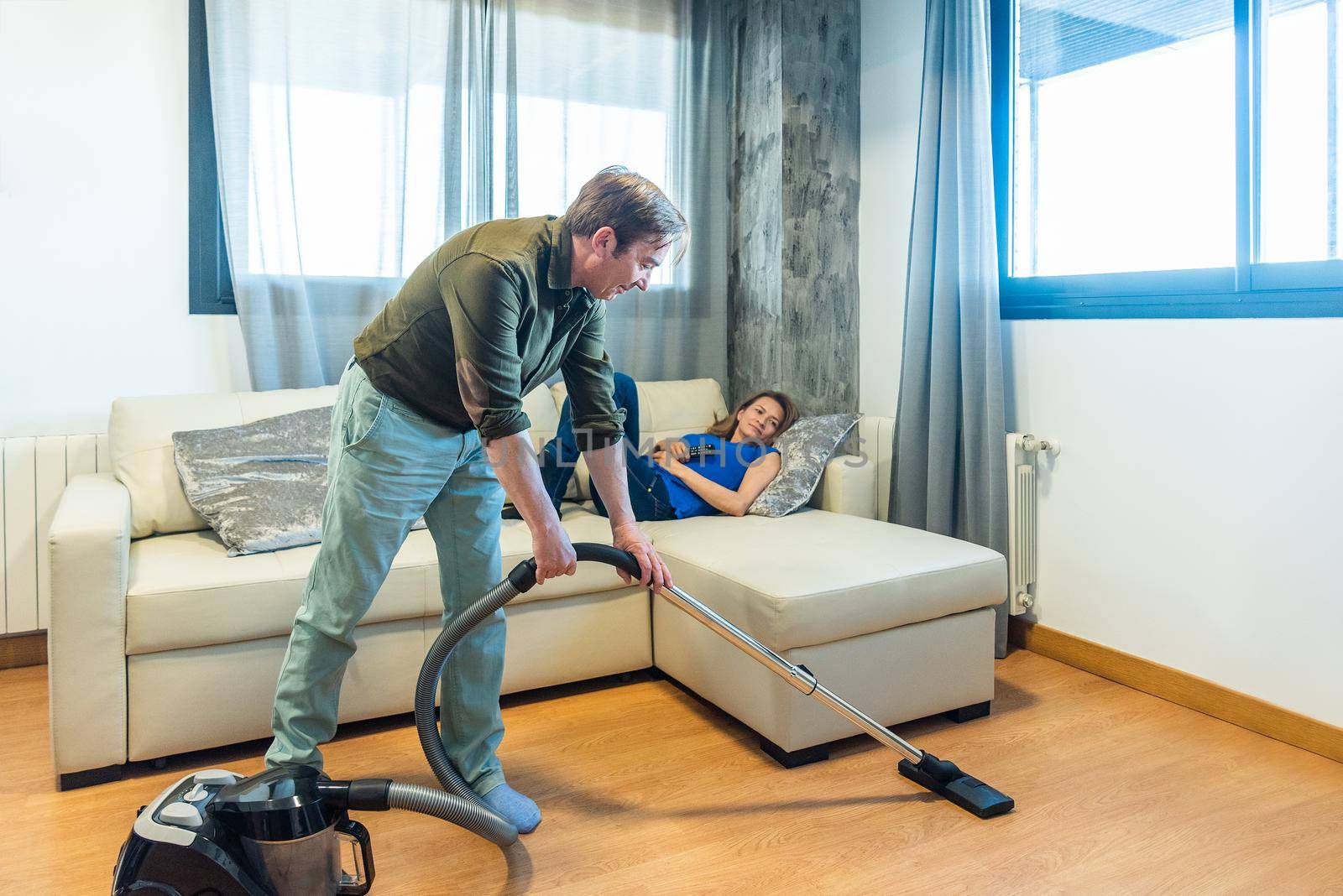 Man vacuuming the living room while his wife is lying on the couch. High quality photo