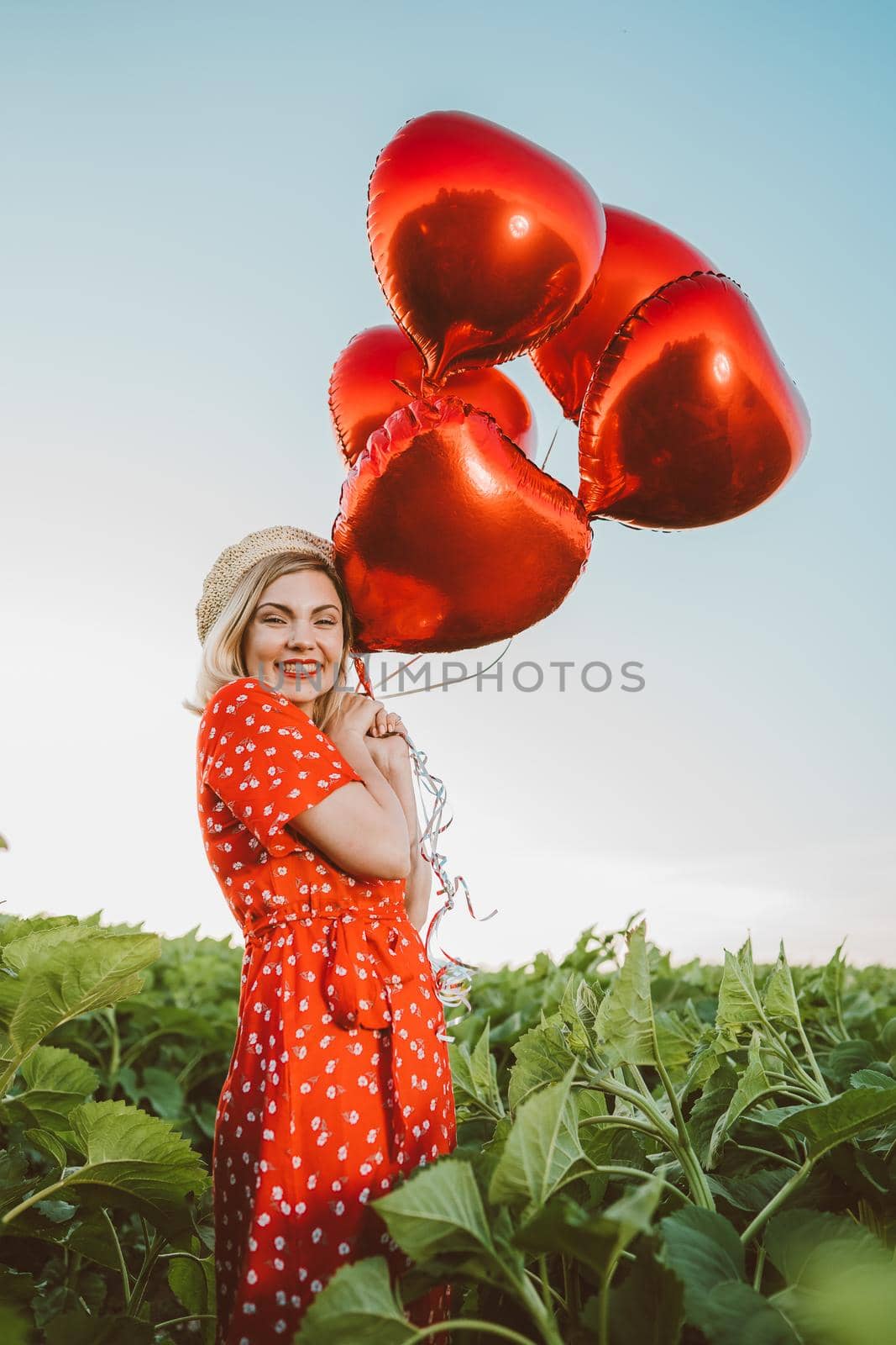 Portrait of attractive woman in red dress posing with heart-shaped balloons on green nature background. Girl in straw vintage hat or beret. Birthday, holiday, celebrate freedom concept. High quality
