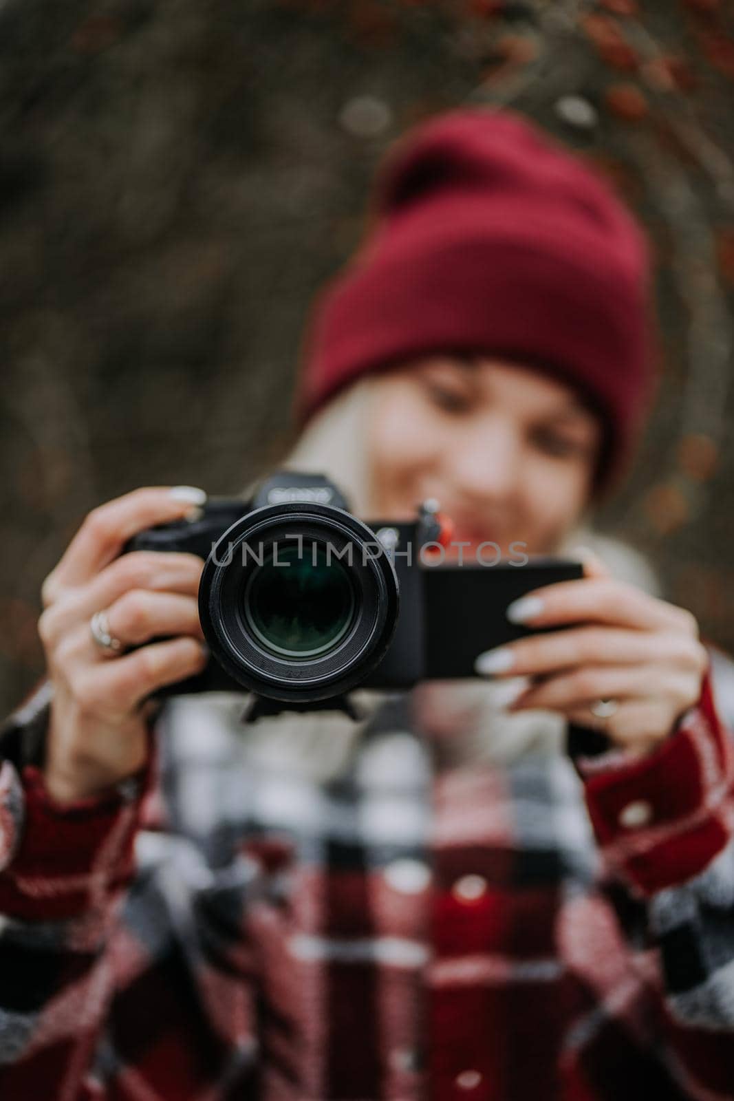 Portrait of young woman using camera in forest. Girl filming and smiling, lady in trendy shirt and red beanie hat. by kristina_kokhanova