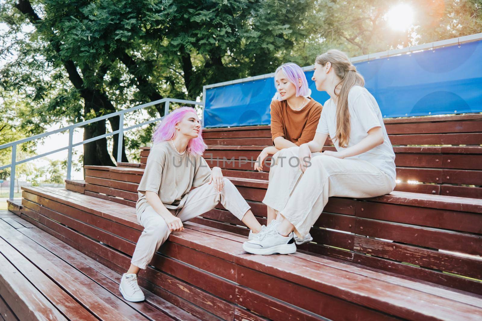 Modern teenage girls with colorful dyed hair sitting on bench in park. Women chatting, gossiping and laughing. Friendship concept. by kristina_kokhanova