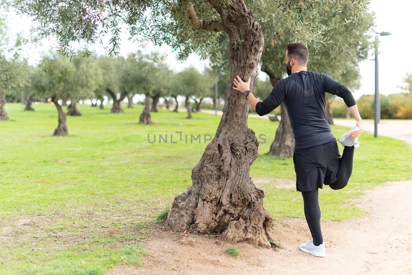 Young runner man with mask stretching in a park leaning against a tree. High quality photo