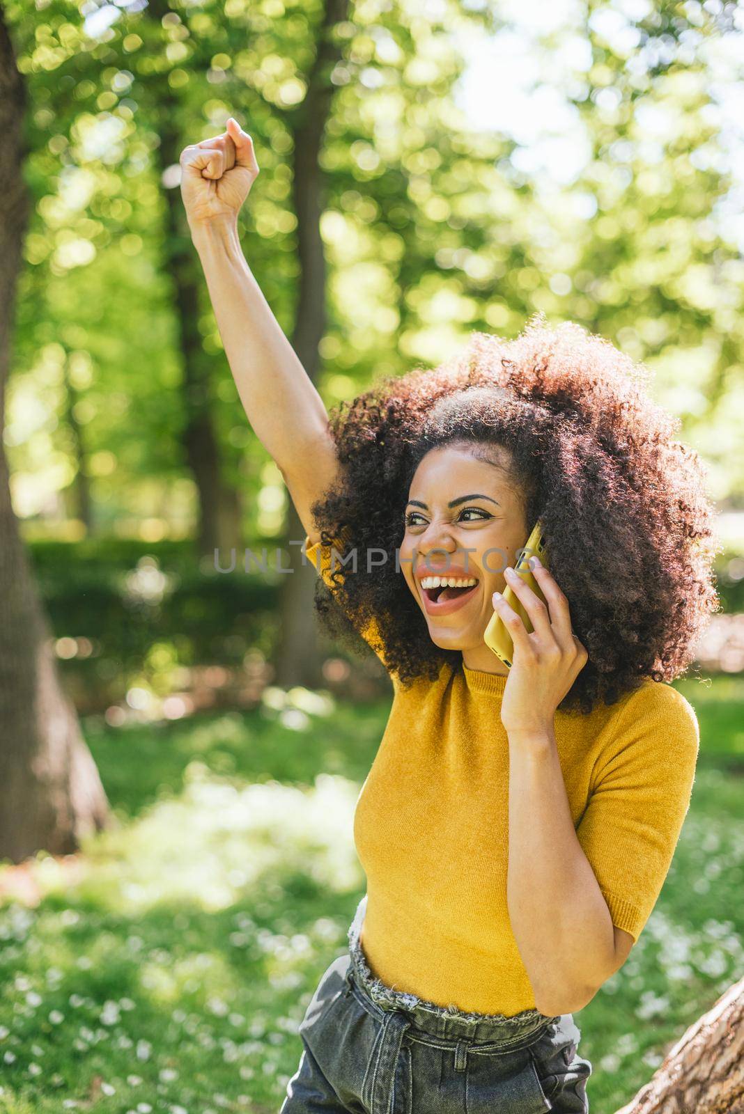 Pretty afro woman talking on the mobile celebrating something with her fist raised, in a garden. Selective focus.