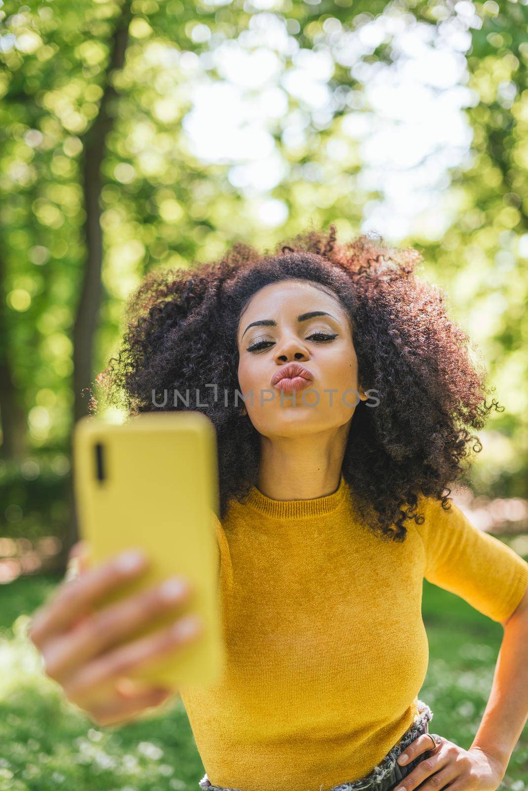 Pretty afro woman taking a selfie blowing a kiss, in the forest. Selective focus.