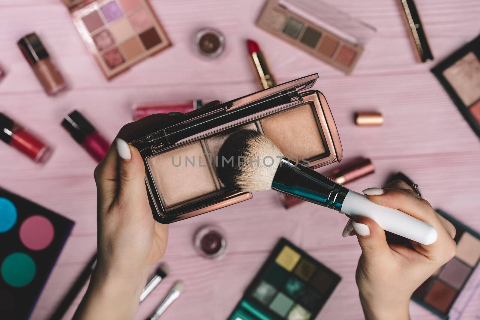 Woman working with face contouring palette - powder, bronzer and highlighter on pink flat lay cosmetics collection background. Tools in beauty industry - lipsticks, eyeshadows, glosses. by kristina_kokhanova