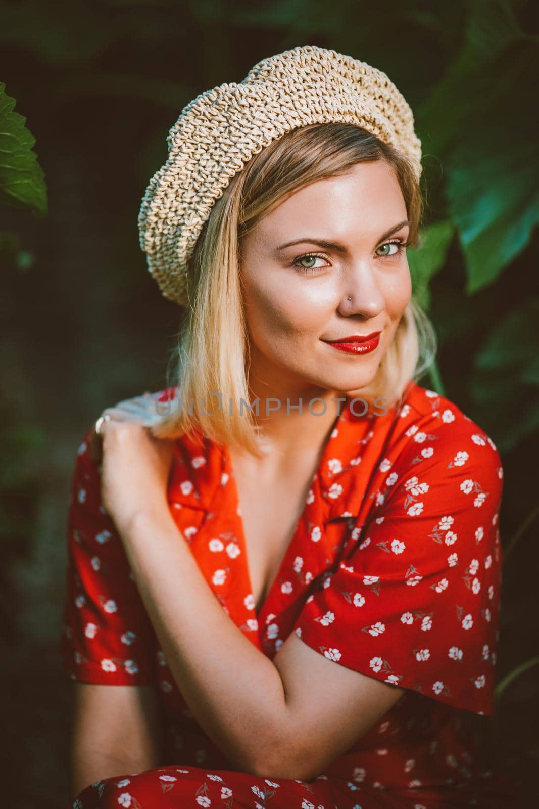 Beautiful portrait of attractive blonde woman in straw beret hat . Girl sitting between plants. Nature background. Retro floral dress, red lips. Pleasant feminine appearance, kind smile. .