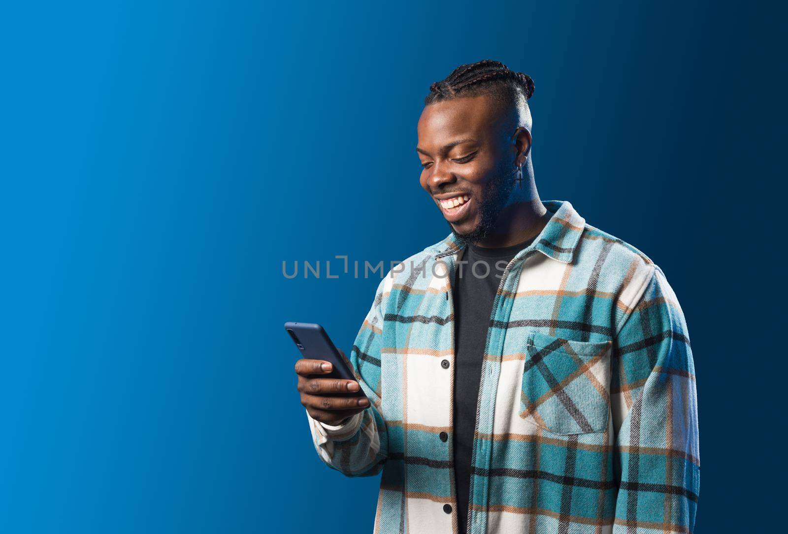 Handsome black man looking at mobile smiling. Mid shot. Blue background.