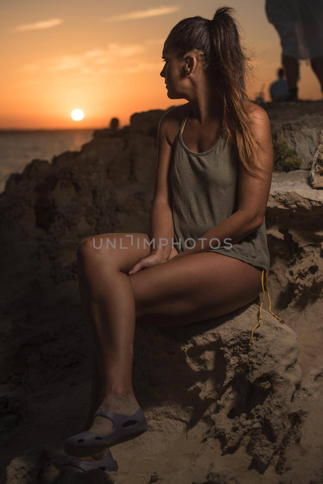 Pretty woman in a T-shirt and swimsuit, looking at a beautiful sunset by the sea, sitting on a rock. Long shot. Formentera island, Spain.