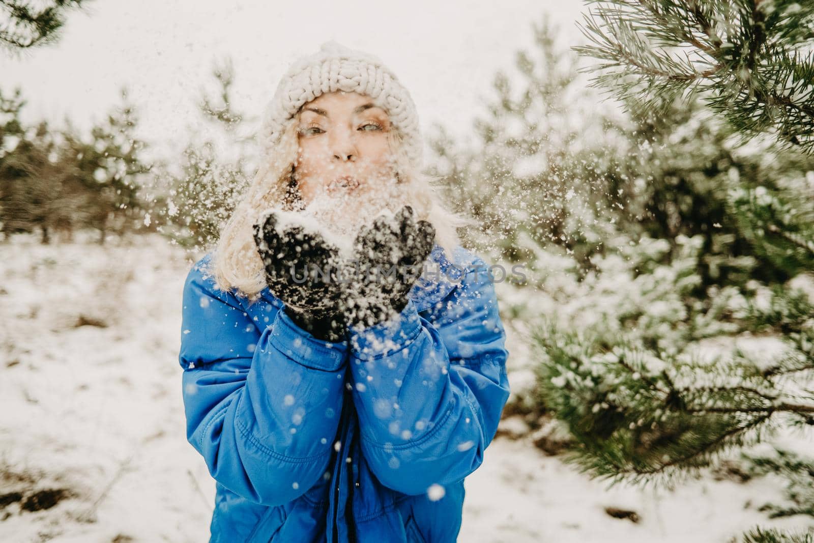 Young cute girl blowing on first snow from hands to camera and then scatters it to sides. Winter forest background. Portrait of cute woman childishly rejoices nature. High quality