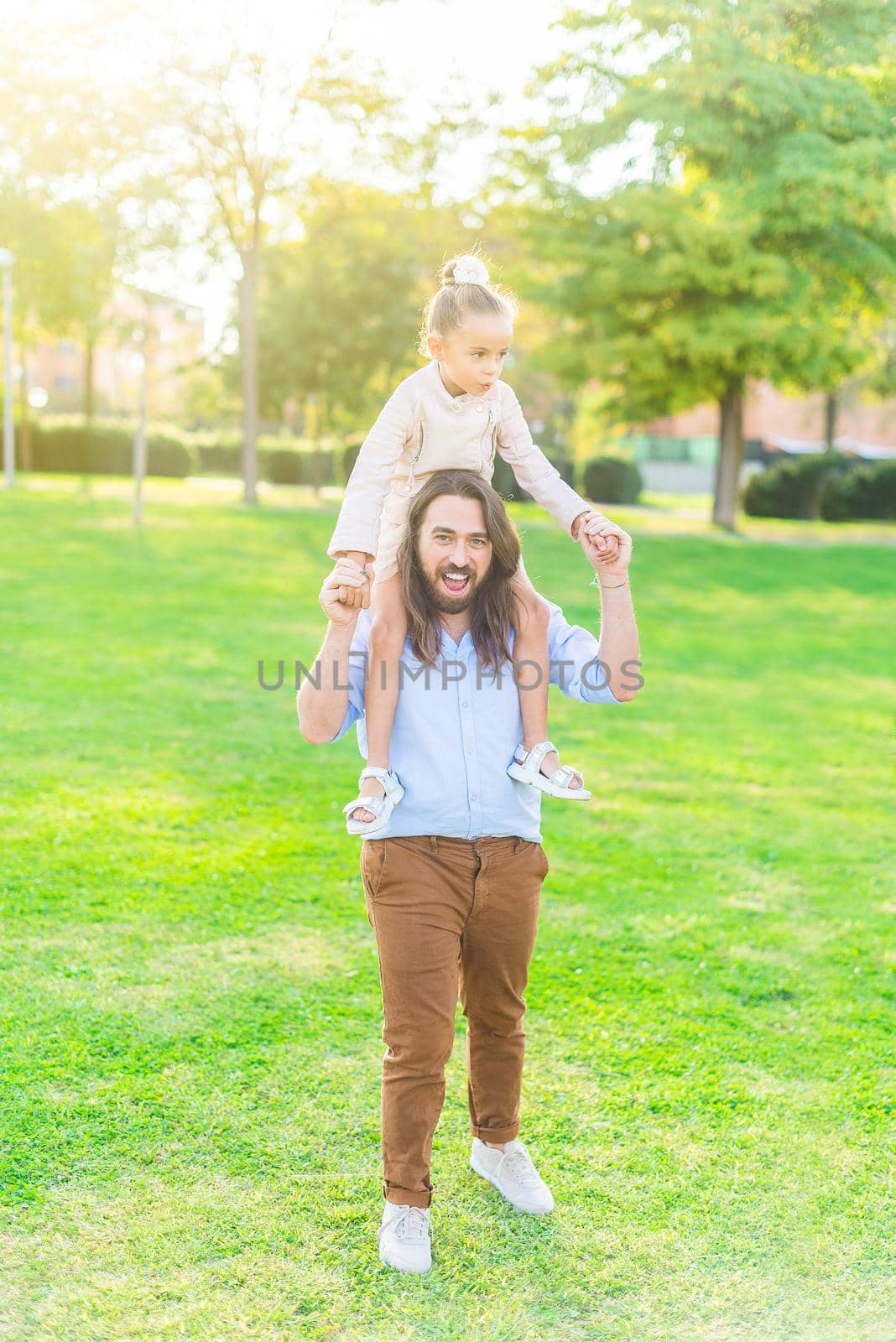 Vertical image of a enthusiastic young father holding her little daughter on his shoulder in park