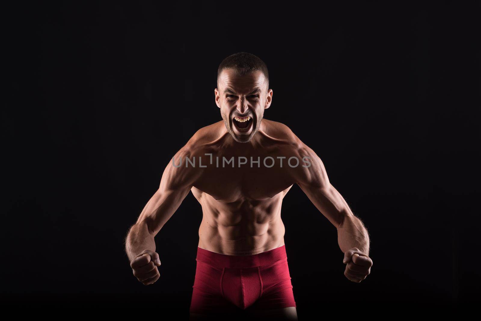 Furious shirtless man showing his muscles. Mid shot. Black background.