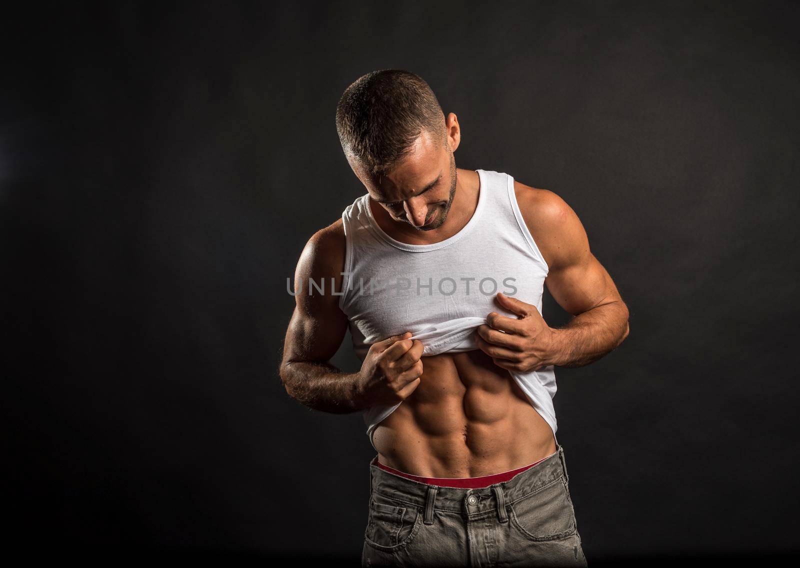 Athletic man lifting his shirt to show his abs. Mid shot. Black background.