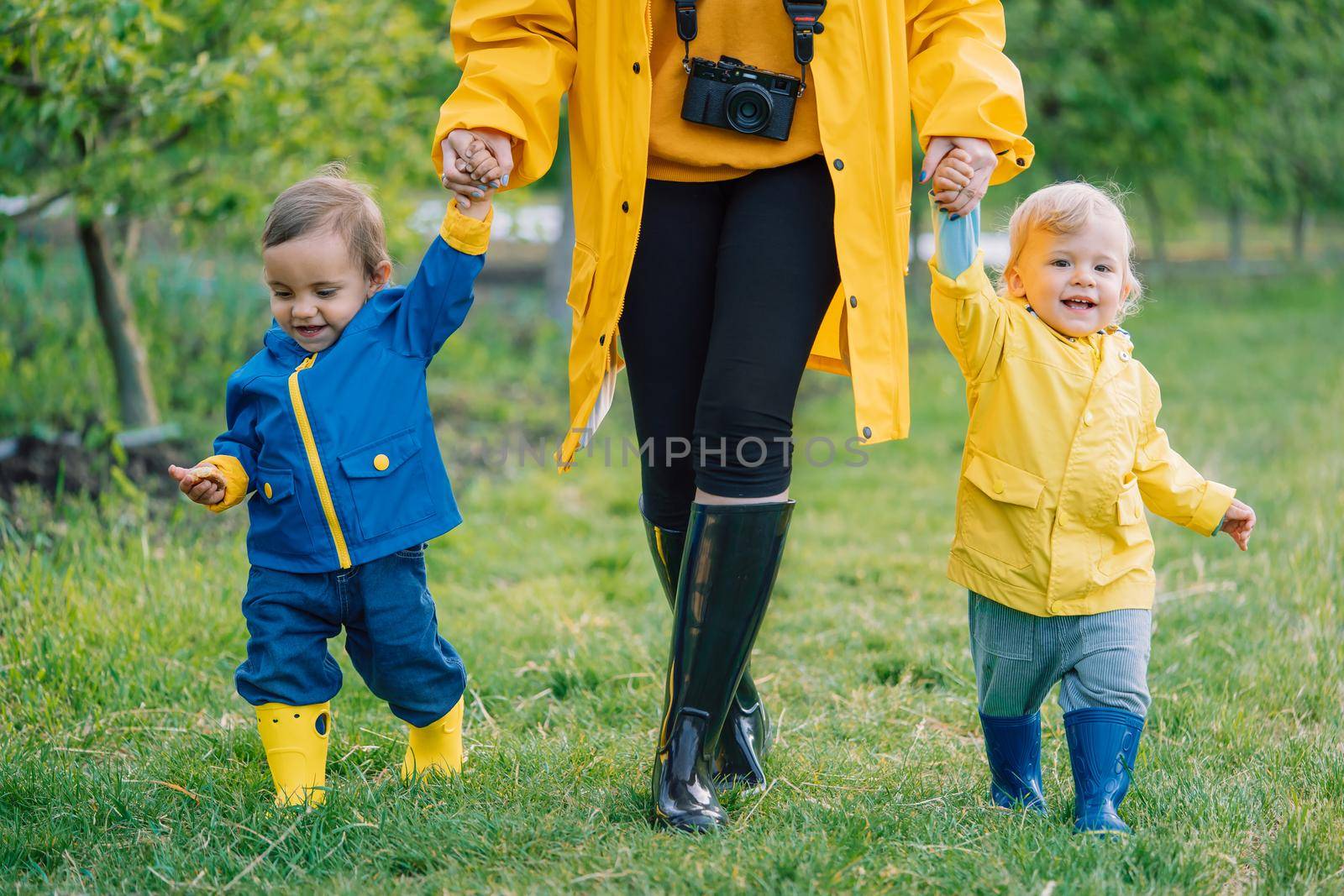 Mother and cute babies twins in colorful raincoats walking in green forest after rain. Children holds mom's hand. Love, care, attachment, family, kids concept. High quality photo