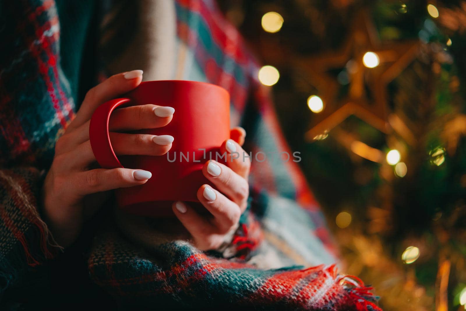 Unrecognizable woman holding cozy red mug on glowing Christmas tree background. Girl with hot drink - tea, coffee or cocoa. Concept of the New Year, comfort and warmth. by kristina_kokhanova