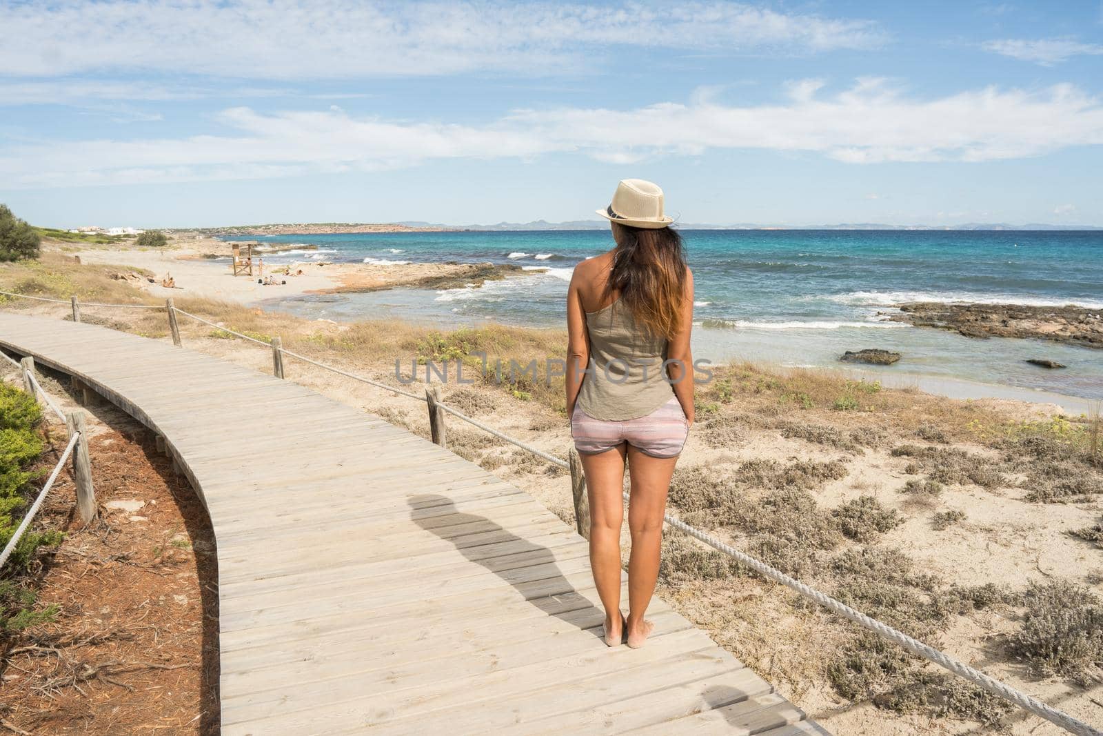 Woman in a T-shirt and hat, standing looking out to sea from a little path made of wooden boards. Long shot. Formentera island, Spain.