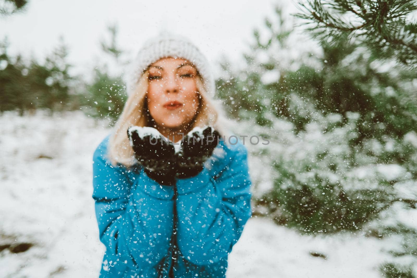 Happy woman blowing snow from palms with mittens to camera on winter forest background. Portrait of cute girl childishly rejoices first snow. by kristina_kokhanova
