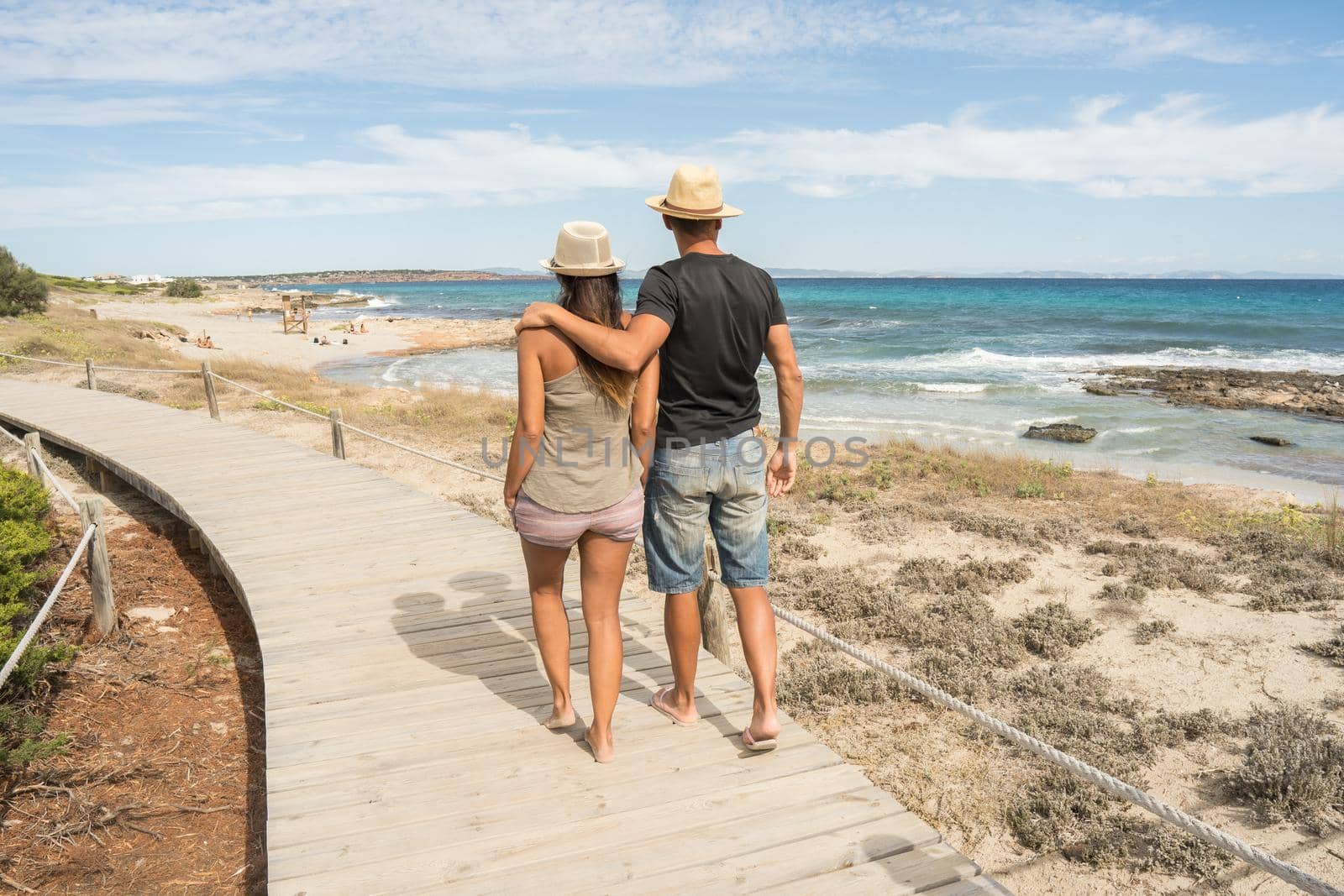 Couple of man and woman in love walking along a path of wooden boards by the sea. Long shot. Formentera island, Spain.