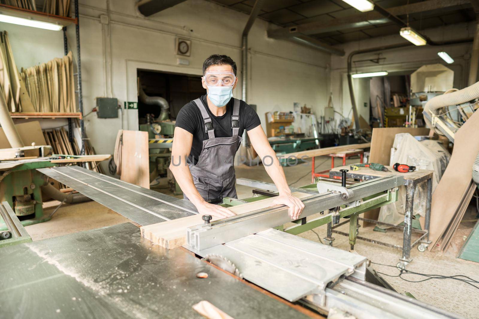 Worker with mask and protective equipment cutting on a sliding table saw in a factory. Mid shot.