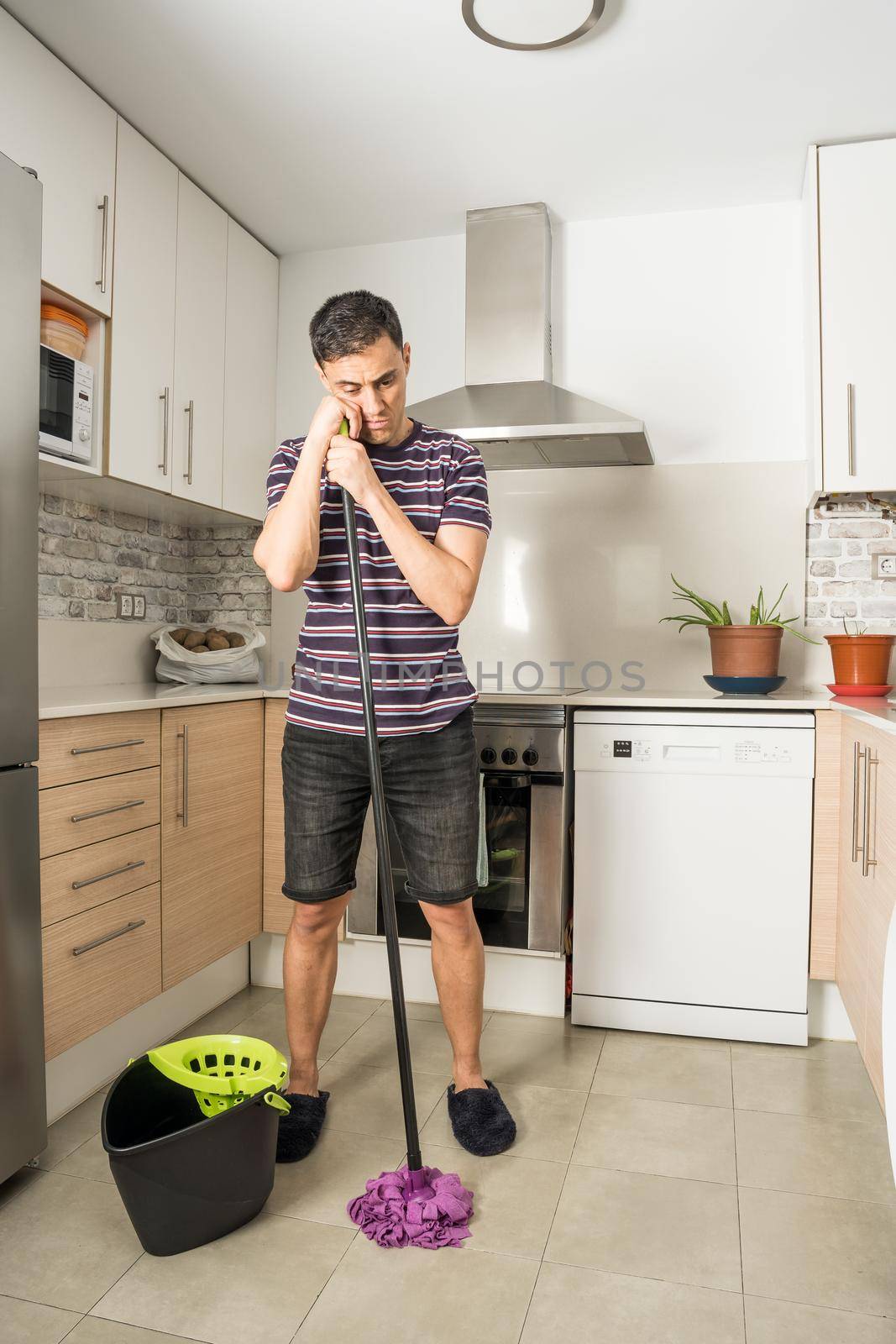 Sad and pensive man wearing casual clothes and slippers mopping the kitchen floor. Full body.