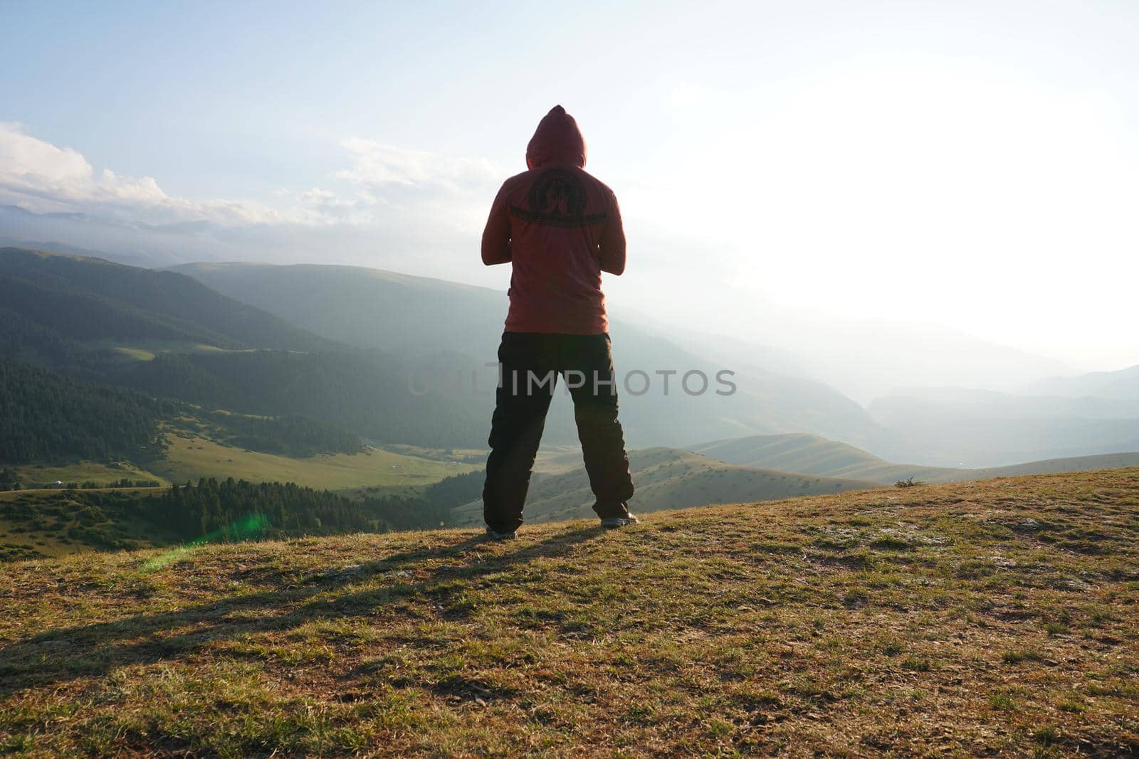 A guy on the edge of the hill admires the view. In the distance you can see high mountains and green hills covered with forest. Wide margins. Clouds are visible and sun is shining brightly