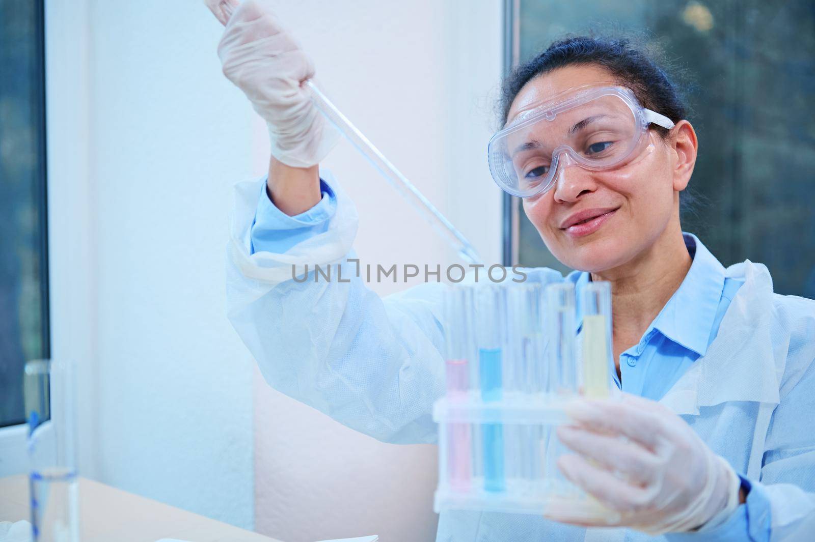 Inspired Latin American woman, lab assistant, scientist chemist using a glass dropper, dripping few chemical reagent into test tubes, while conducting experiment in a clinical research laboratory