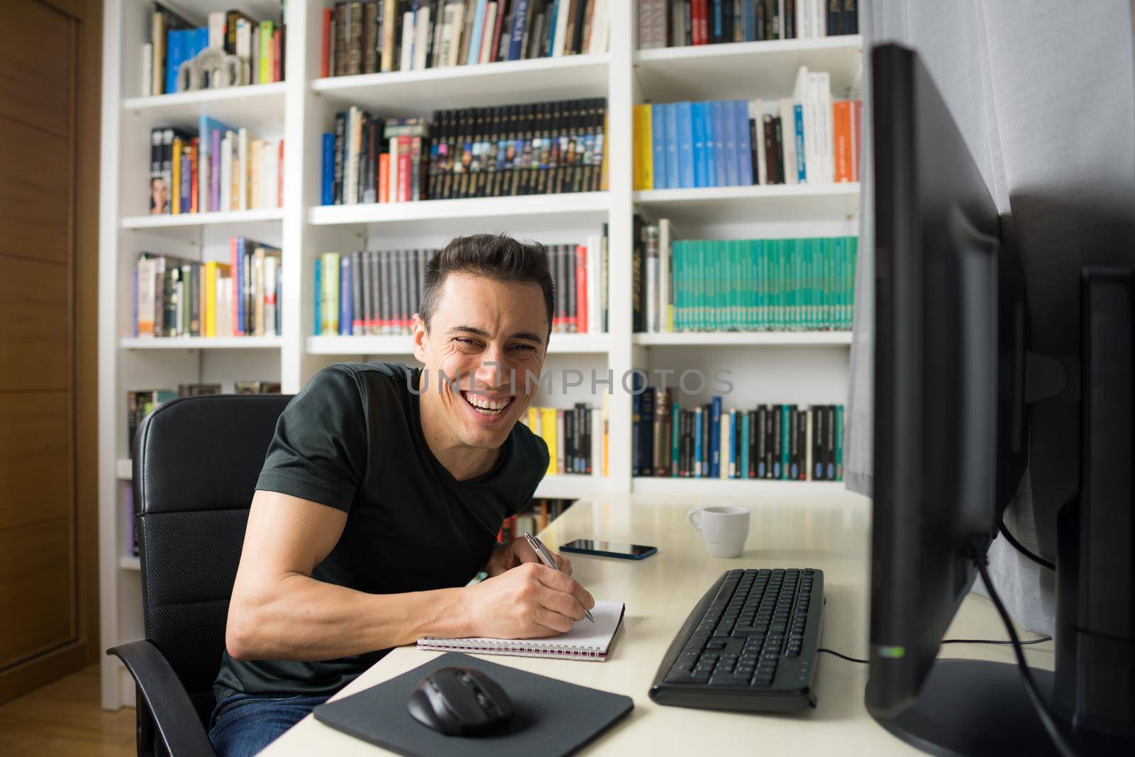 Smiling and happy man in his room sitting at the computer looking at camera, wearing a shirt. Mid shot.