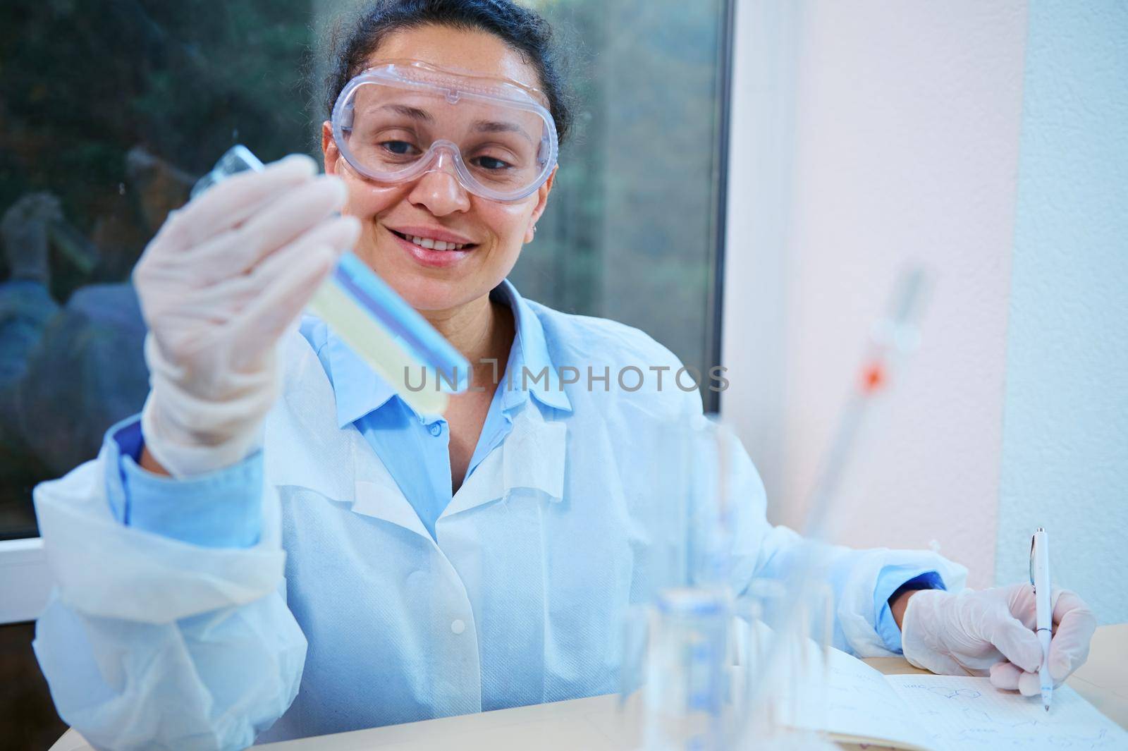 Pleasant African American woman, lab technician, scientist, pharmaceutical chemist holding test tubes with chemical substances, testing the scientific experiment in a clinical research laboratory.