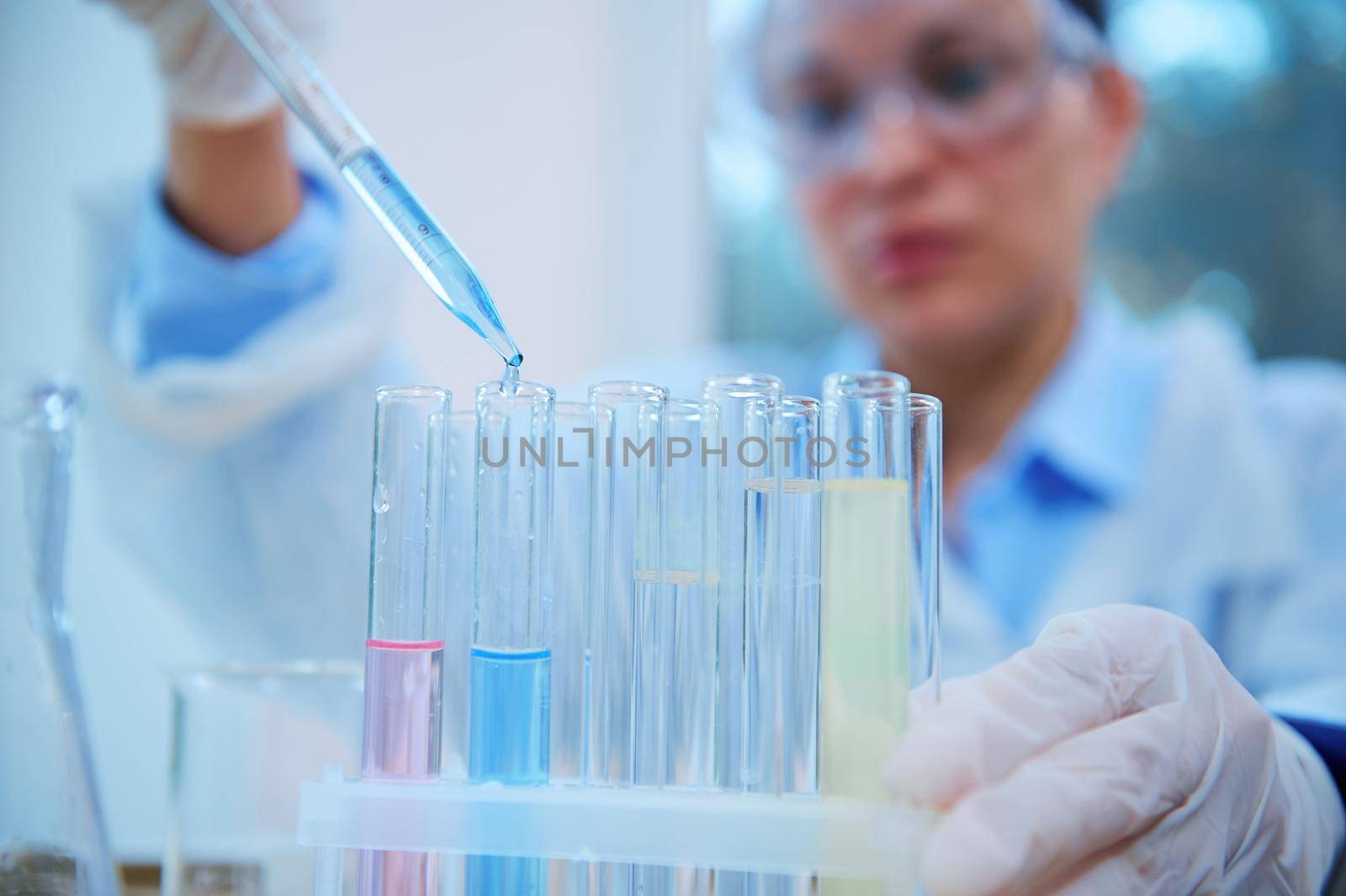 Selective focus on a drop of a blue liquid substance, dripping from a glass laboratory pipette and test tube on a tripod, over blurred scientist conducting experiment in a clinical research laboratory