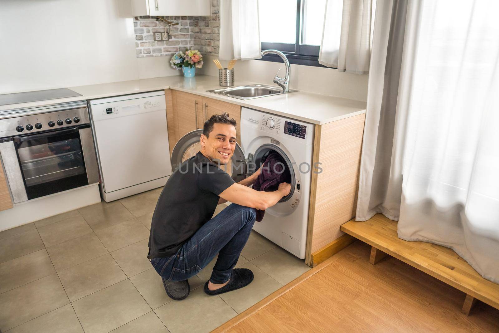 Crouching man putting clothes into the washing machine in the kitchen. Full body.