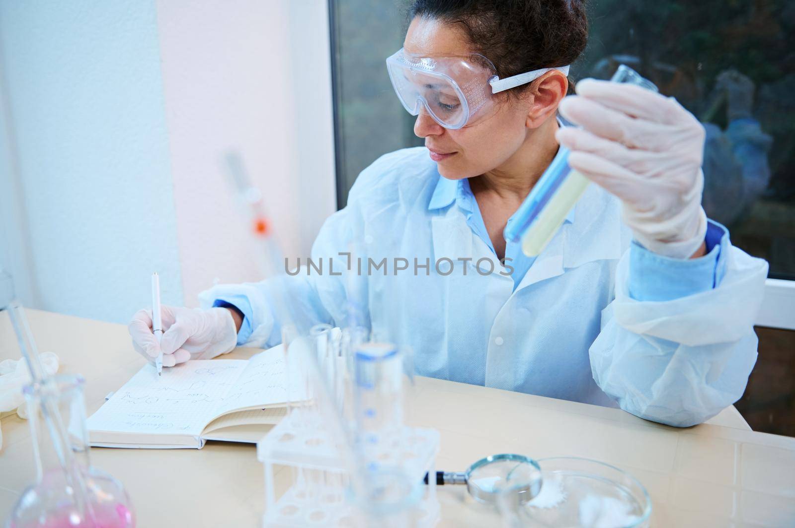 Multiethnic woman scientist, pharmacologist, researcher in a white lab coat and goggles writes in a notebook the results of experiments carried out in test tubes in a clinical research laboratory