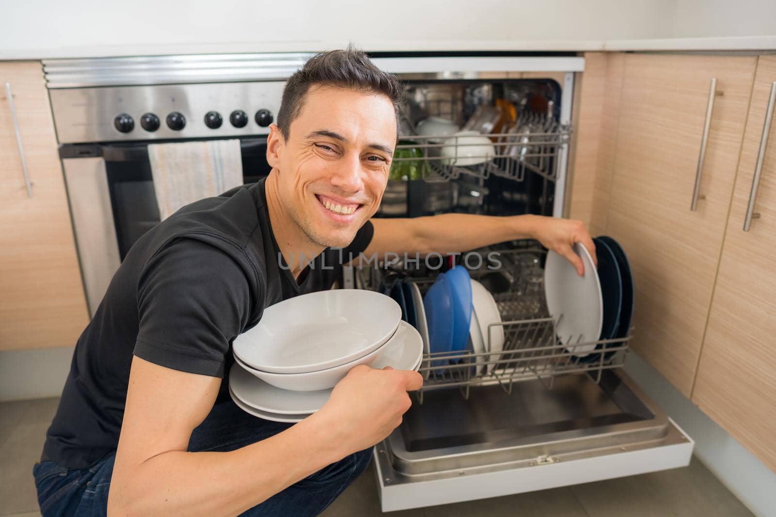 Man taking dishes out of the dishwasher in the kitchen and looking at camera smiling. Close up.