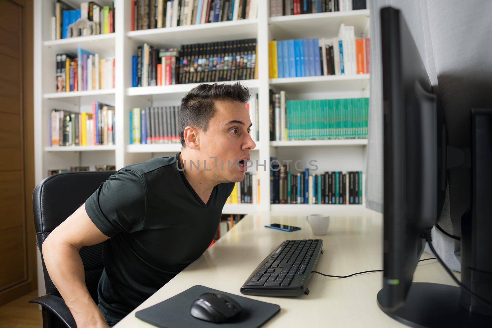 Seated man in black shirt looking at the computer screen very surprised. Mid shot.