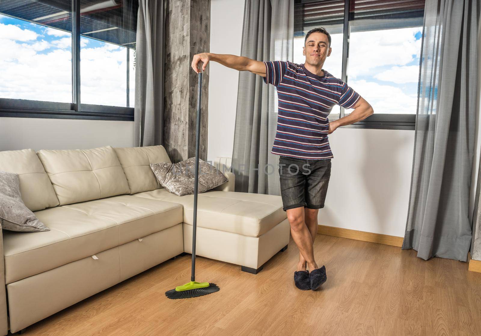 Smiling man in casual clothes and slippers sweeping his living room. Full body.