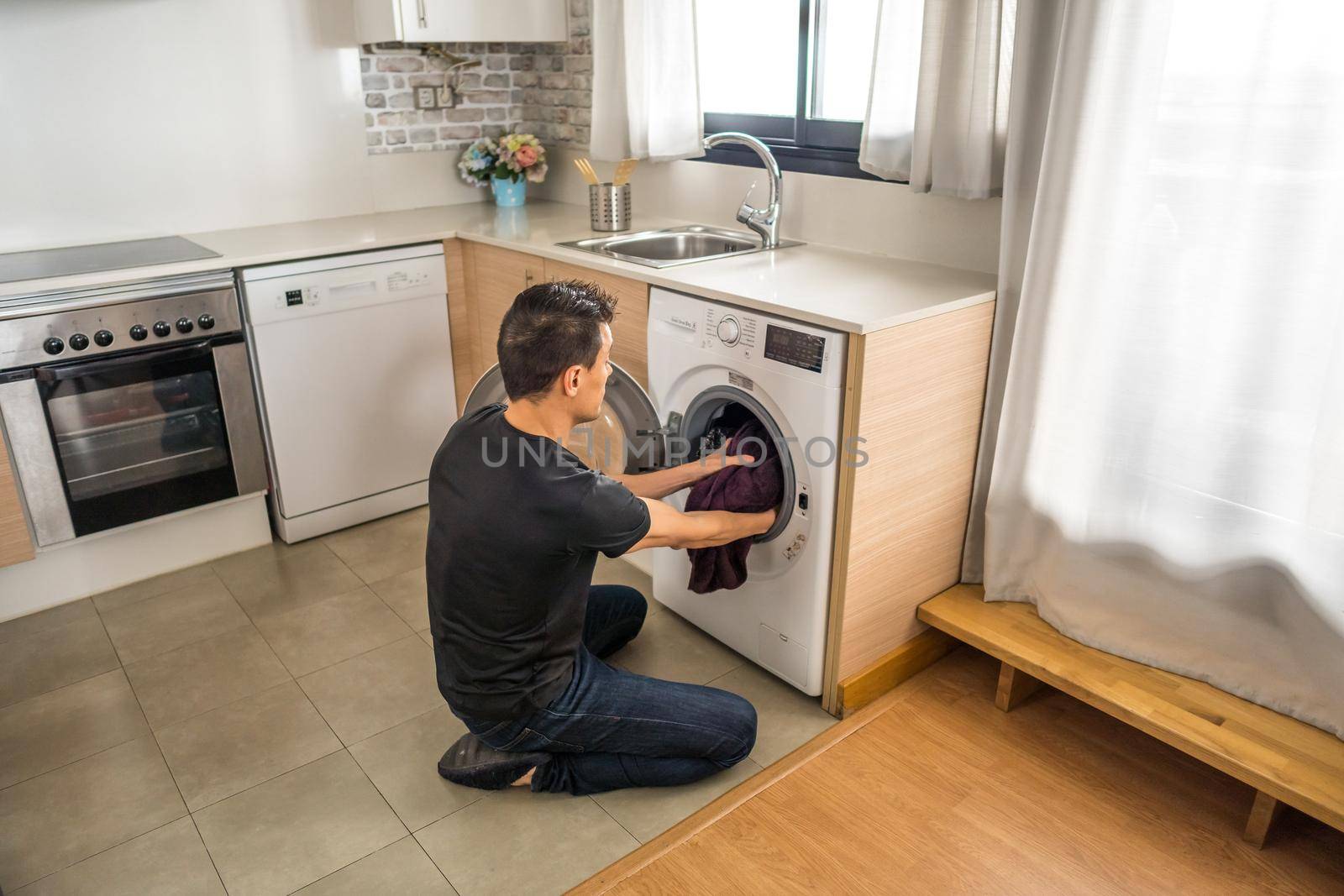 Crouching man putting clothes into the washing machine in the kitchen. Full body.