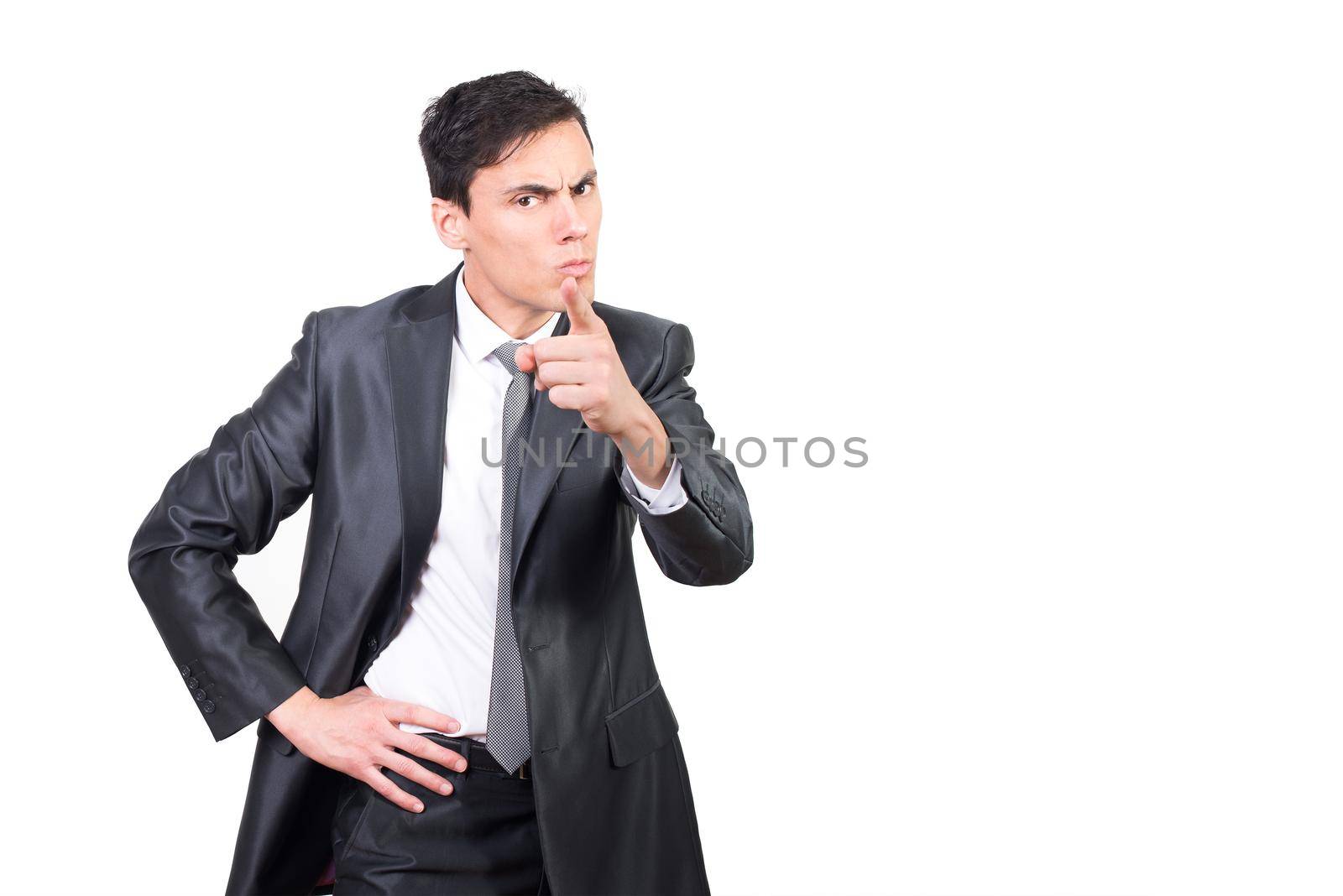 Angry young businessman in classy suit pointing finger at camera while standing with hand on waist against white background