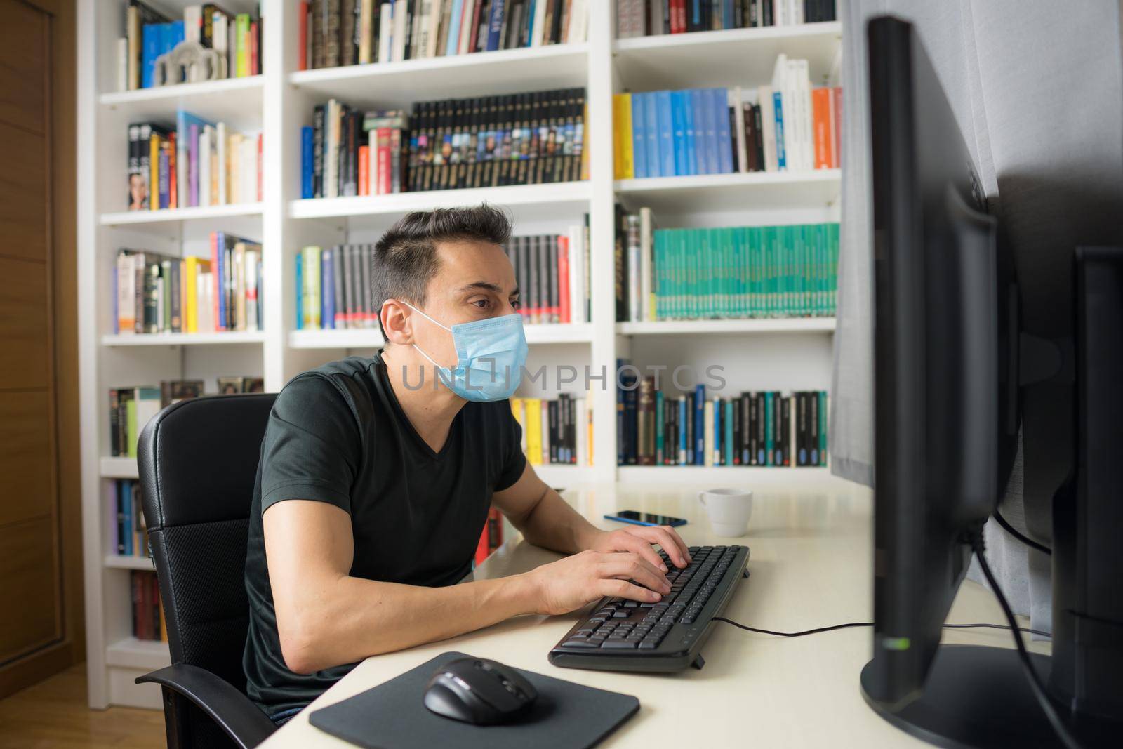 Man teleworking at home, sitting in front of the computer wearing a shirt and mask. Mid shot.