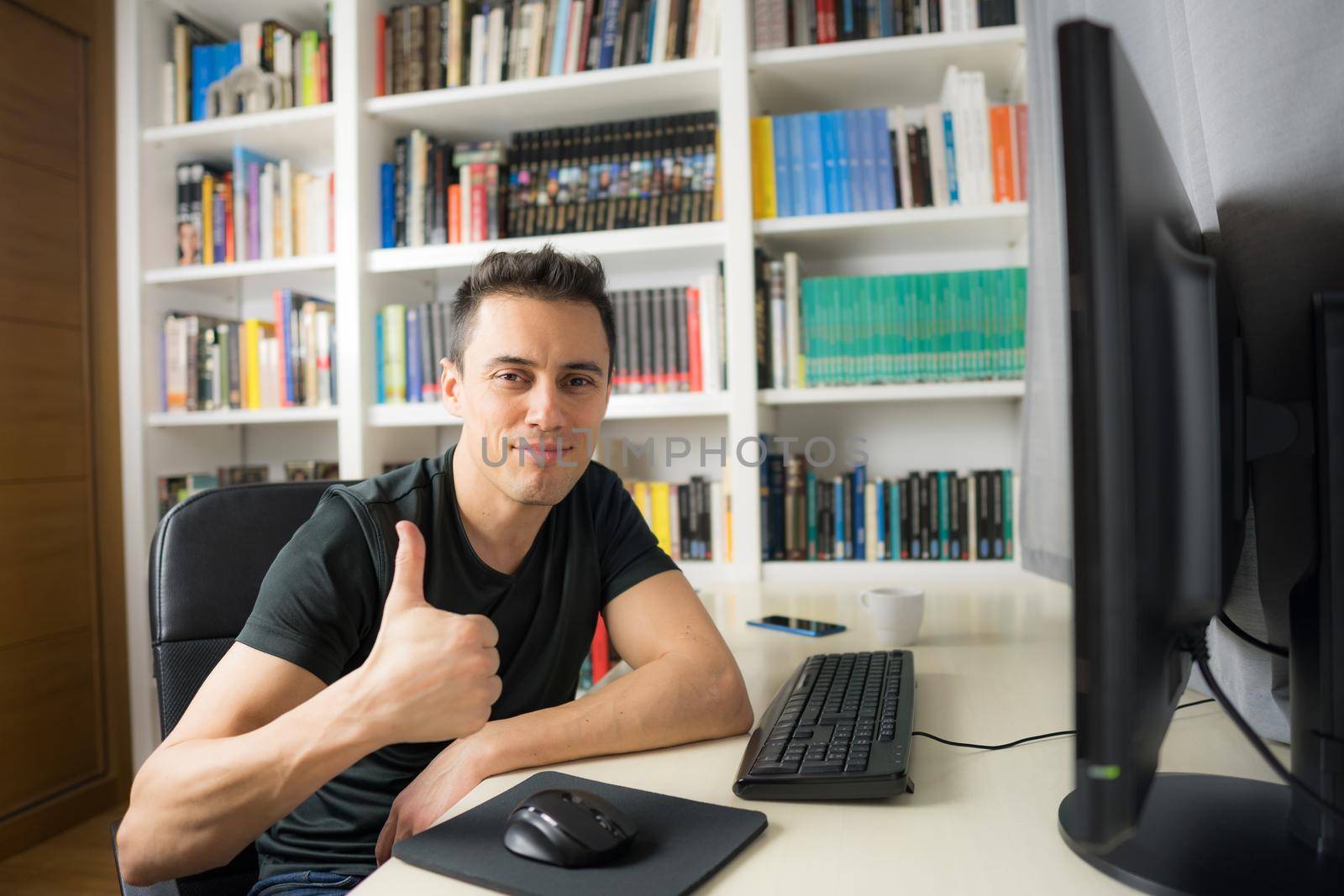 Smiling and happy man in his room sitting at the computer looking at camera, wearing a shirt. Mid shot.
