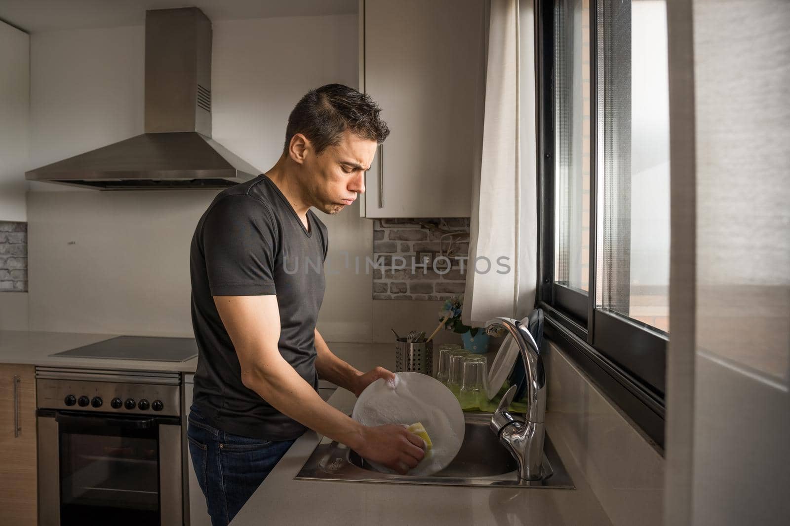 Tired man washing dishes in the kitchen next to a window. Medium shot.
