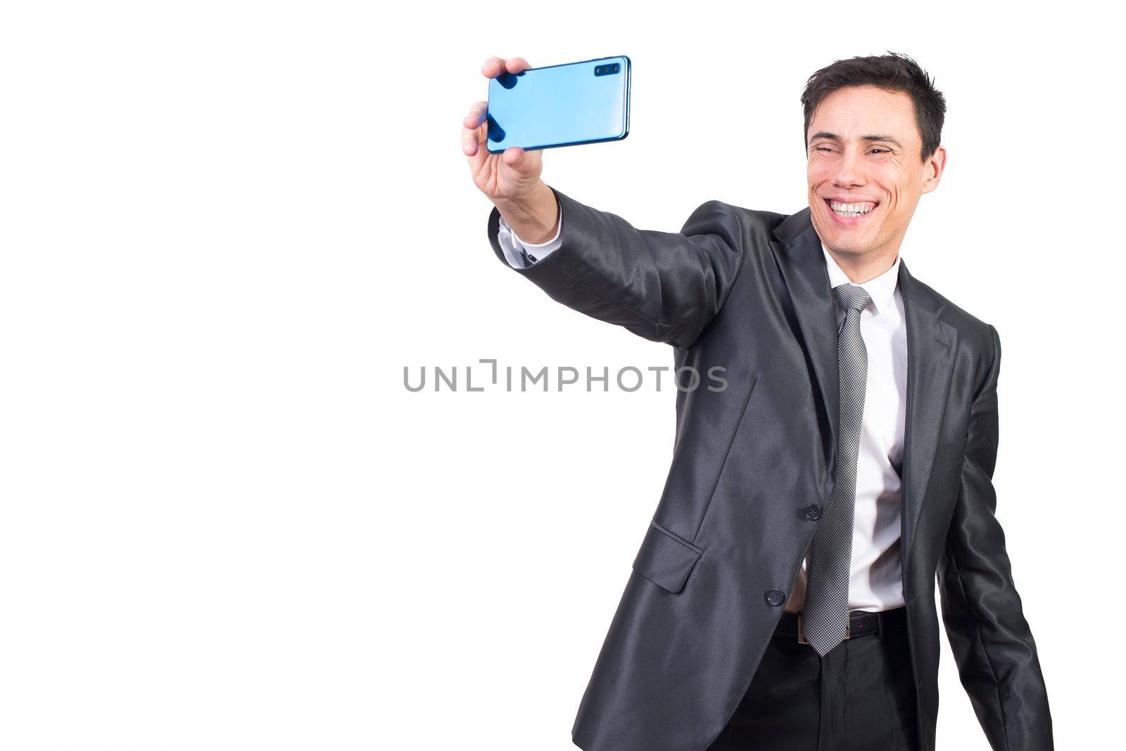 Happy male in formal suit smiling while taking self portrait on modern smartphone while standing isolated on white background in studio