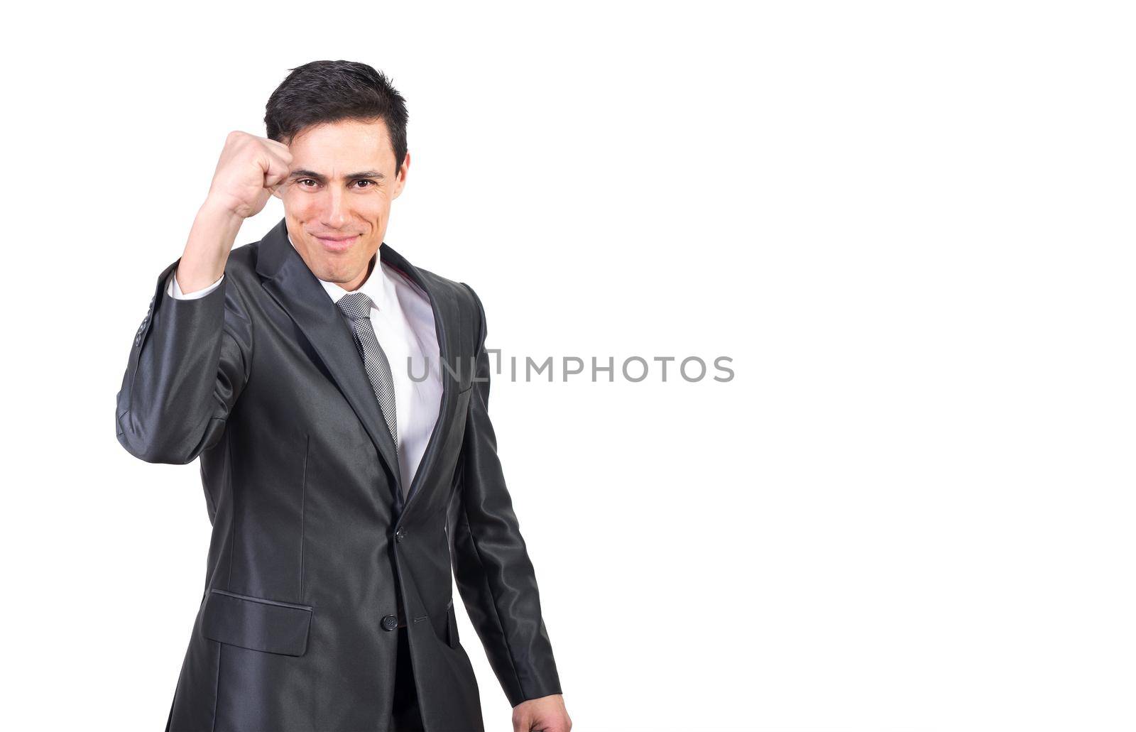 Self assured young male entrepreneur with dark hair in formal suit and tie showing fist and smiling at camera while standing against white background in studio