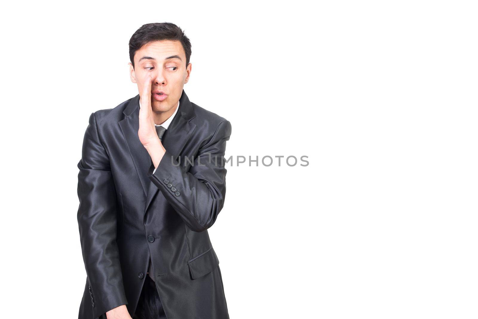 Positive man in elegant costume showing gesture with hand imitating whispering secret standing against white background