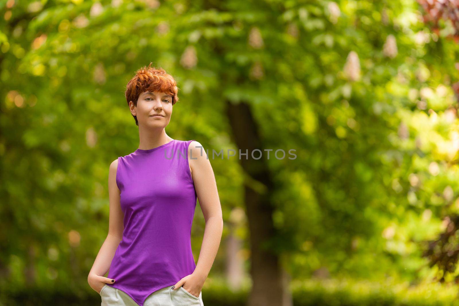 Self assured androgynous woman in casual clothes holding hands in pockets and looking at camera while spending summer day on blurred background of garden