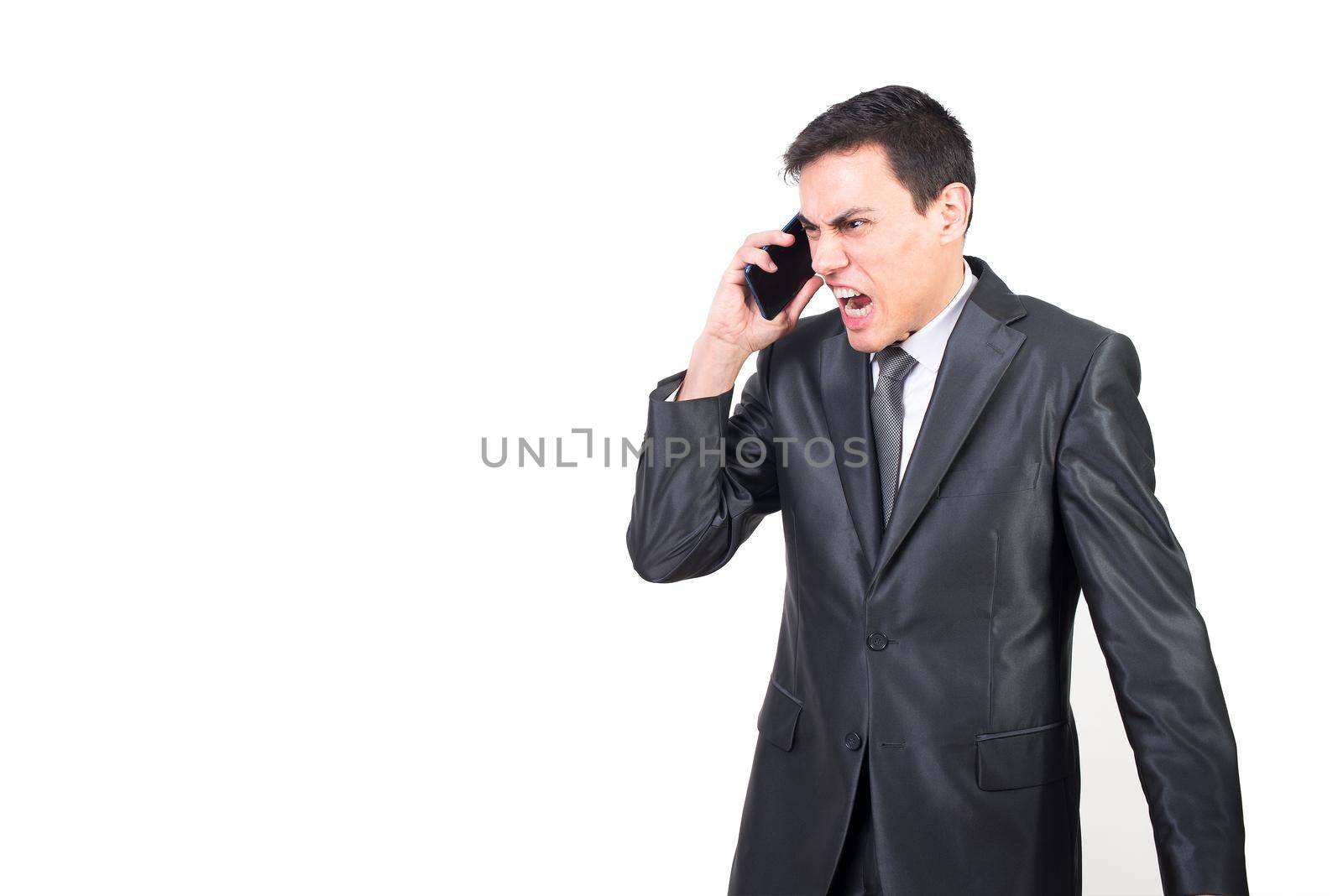 Serious angry businessman in formal suit talking on mobile phone while standing against white background in studio