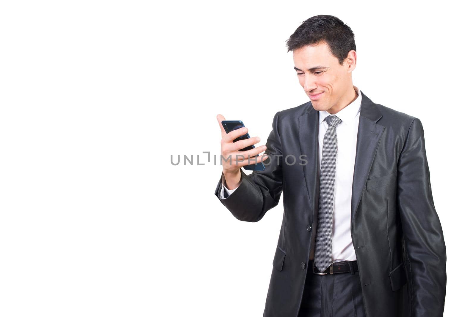 Positive male in elegant suit watching video on cellphone with smile while standing isolated on white background in light studio