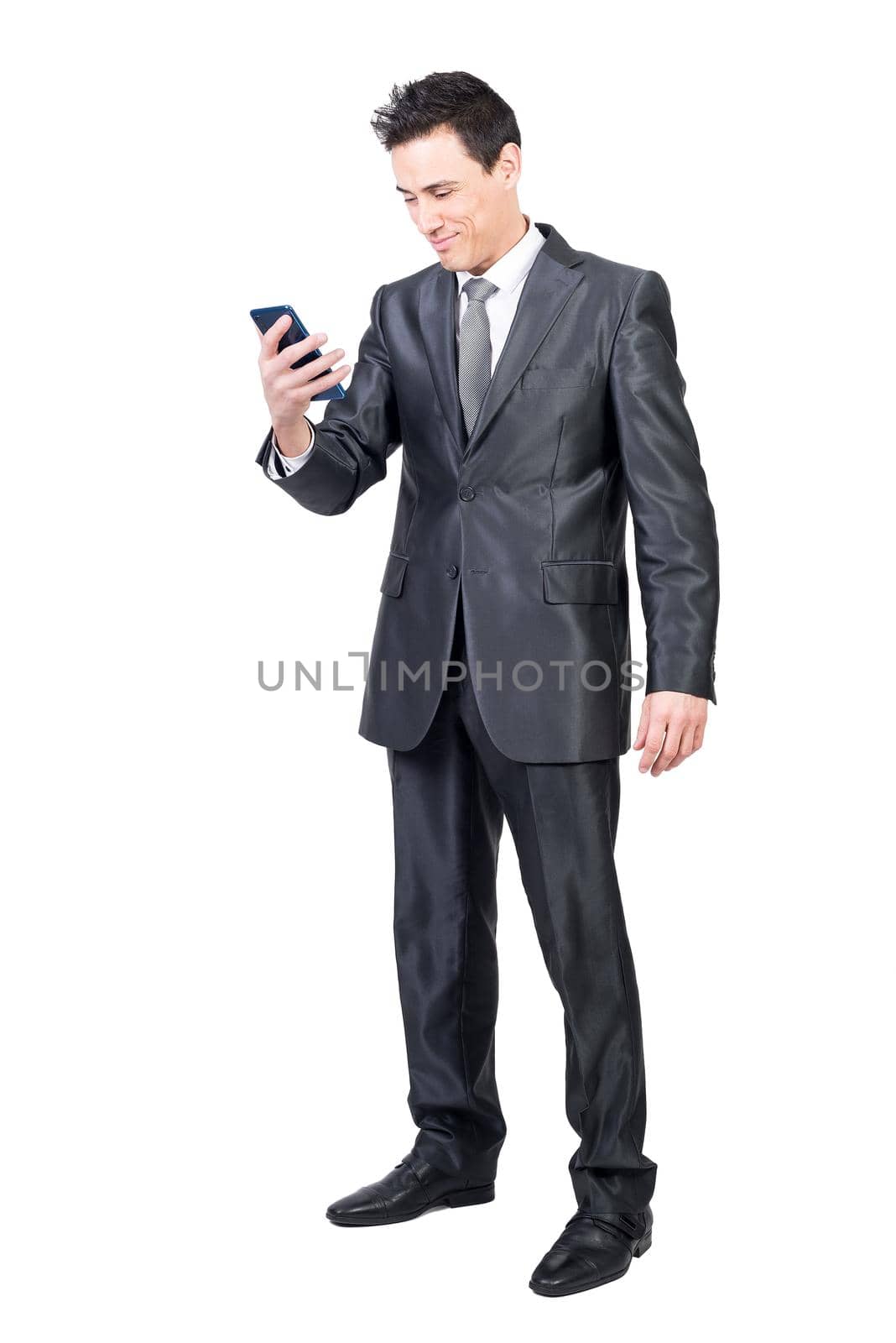 Full body of glad male in elegant suit surfing internet on modern cellphone isolated on white background in light studio