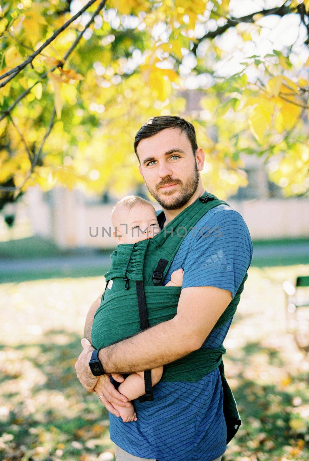 Dad hugging a baby in a sling standing under a tree in the park by Nadtochiy