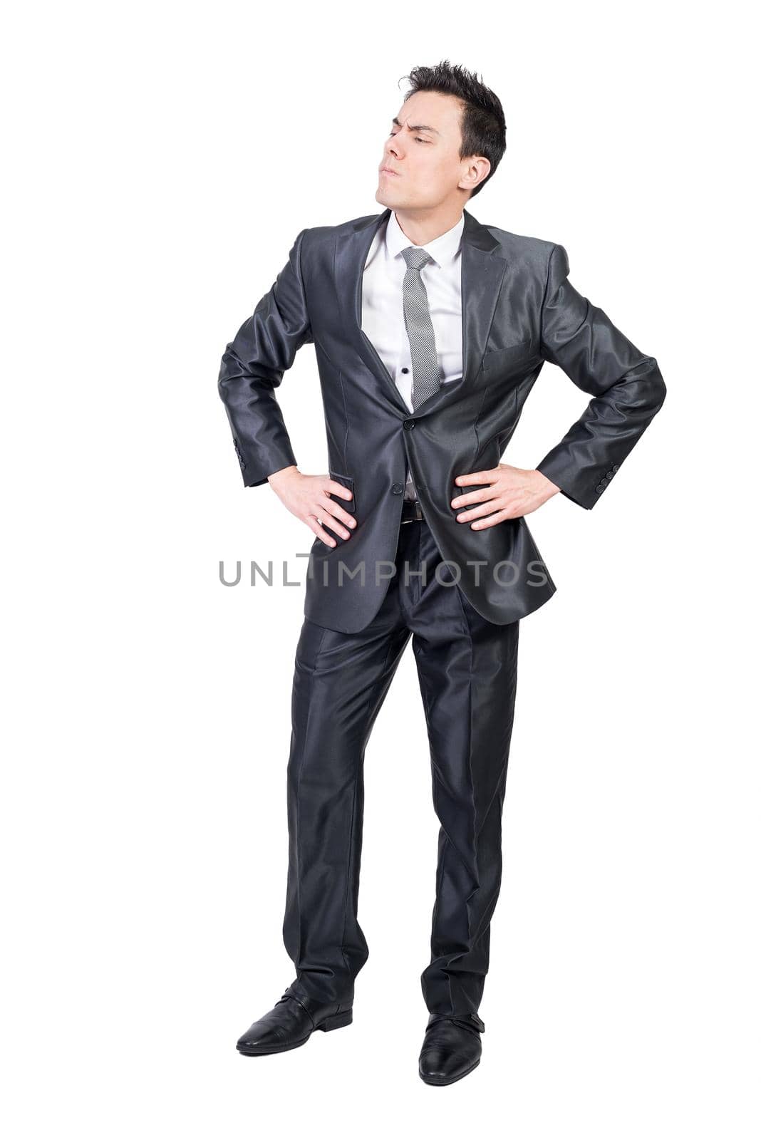 Full body of confident young businessman with dark hair in classy suit and tie standing isolated on white background with hands on waist and looking away with concentration