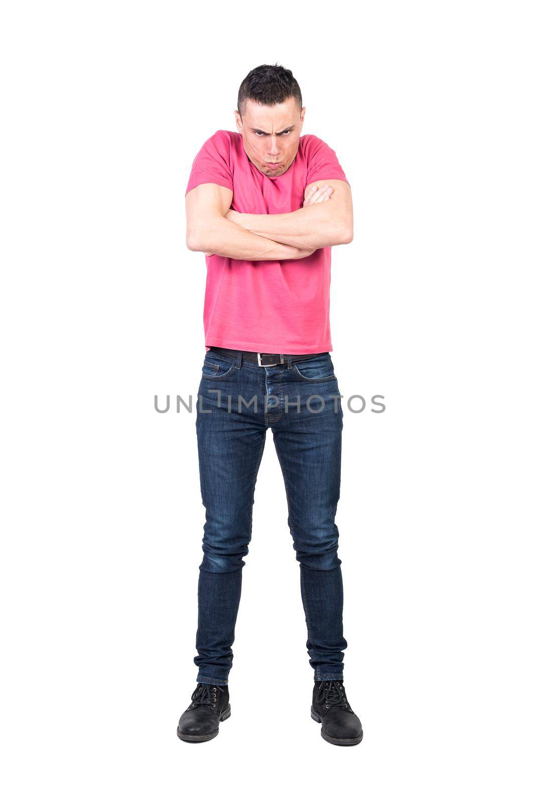 Full body of angry young male with dark hair in pink t shirt and jeans making face and gazing at camera while standing against white background with crossed arms